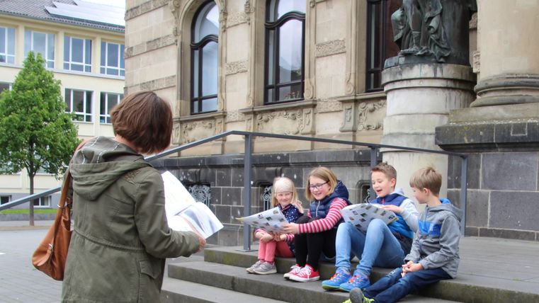 Eine Gruppe von Kindern sitzt auf den Stufen vor einem Gebäude und hört einer Lehrerin zu. Sie halten Bücher in den Händen und scheinen aktiv zu lernen.