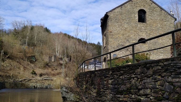 Ein altes Steingebäude neben einem ruhigen Gewässer. Im Hintergrund sind kahle Bäume und hügelige Landschaft erkennbar.