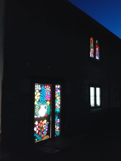 Ein Gebäude mit bunten Glasfenstern, die nachts leuchten. Die Fenster zeigen florale Muster und fügen dem dunklen Umfeld Farbe hinzu.