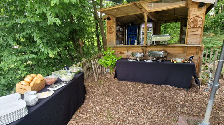 Ein gemütlicher Essensbereich im Freien mit einem Holzstand und einem Buffet. Auf einem Tisch liegen frische Brötchen, Salate und Geschirr, umgeben von Bäumen.