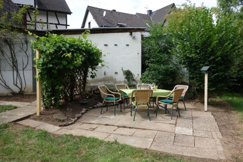 Ein gemütlicher Sitzbereich im Garten mit einem runden Tisch und Stühlen. Umgeben von Pflanzen und einer weißen Wand.