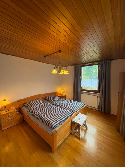 Ein gemütliches Schlafzimmer mit einem Doppelbett und Holzdecke. Es gibt Nachttische, eine Stehlampe und ein Fenster mit Vorhängen.