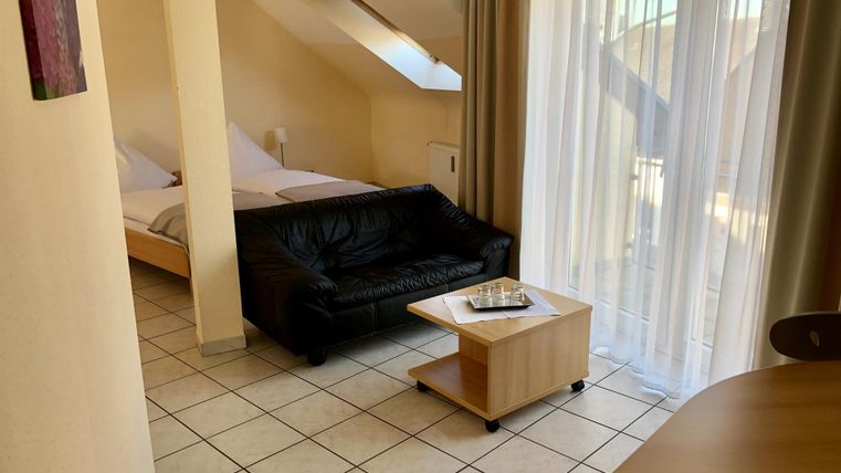 Ein gemütliches Zimmer mit einem schwarzen Sofa, einem Tisch und einem Bett im Hintergrund. Große Fenster lassen viel Tageslicht herein.