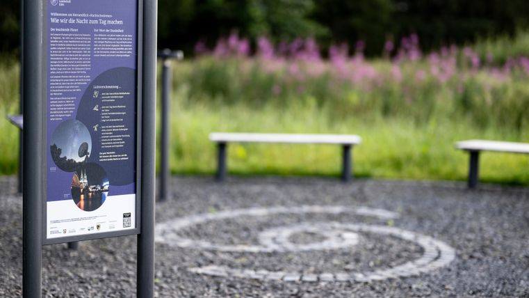 Ein Informationsschild steht auf einem Schotterplatz mit einer spiralförmigen Markierung. Im Hintergrund sind violette Blumen und Sitzbänke zu sehen.