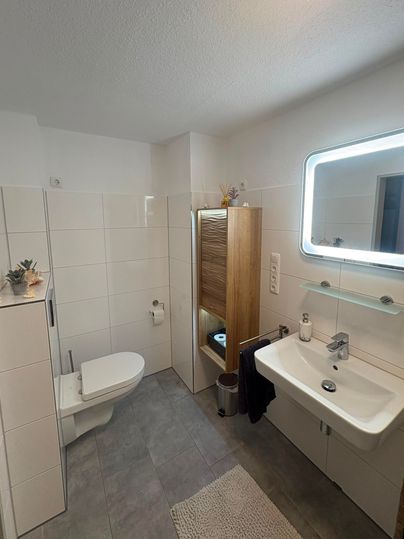Ein modernes Badezimmer mit hellen Fliesen und einer geräumigen Dusche. Es gibt ein Waschbecken, eine Toilette und einen Holzschrank für zusätzlichen Stauraum.