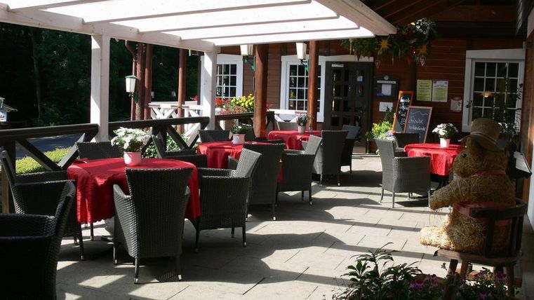 Eine gemütliche Terrasse mit roten Tischen und schwarzen Stühlen. Im Hintergrund ist eine Holzhütte mit Fenstern und Pflanzen zu sehen.