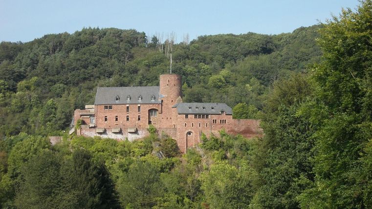 Eine alte Burg, umgeben von grünen Wäldern und Hügeln. Der Himmel ist klar und blau.