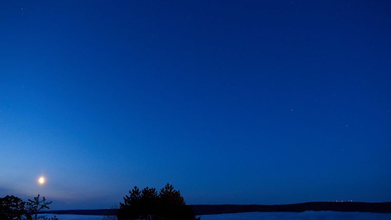Ein klarer Nachthimmel mit einem leuchtenden Mond und einigen Sternen. Unten sind Silhouetten von Bäumen und eine Nebelschicht zu sehen.