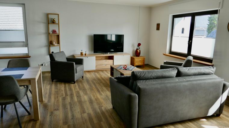 Ein modernes Wohnzimmer mit einer grauen Couch, zwei Sesseln und einem Fernseher. Der Raum hat große Fenster und Holzfußboden.