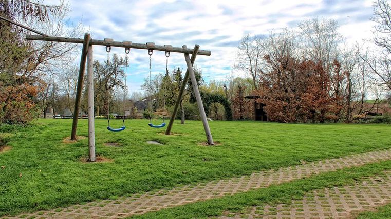 Ein Spielplatz mit zwei Schaukeln steht auf einer grünen Wiese. Im Hintergrund sind Bäume und ein Pavillon zu sehen.