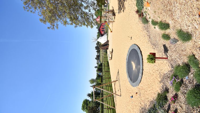Ein Spielplatz mit Sandboden, Schaukeln und einem Trampolin. Im Hintergrund sind Bäume und ein klarer blauer Himmel zu sehen.