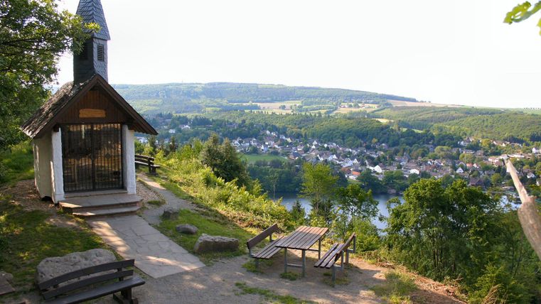 Eine malerische Aussicht von einem Hügel mit einer kleinen Kapelle. Im Hintergrund erstreckt sich ein Dorf neben einem Fluss umgeben von grünen Hügeln.