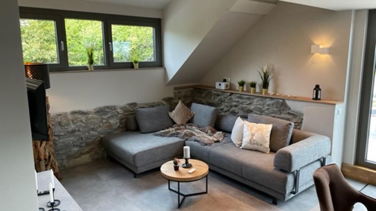 Ein modernes Wohnzimmer mit einer grauen Couch und einem runden Tisch. Große Fenster bieten viel Licht und einen Blick auf die grüne Umgebung.