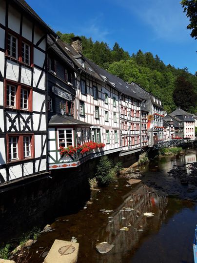Ein malerisches Fachwerkhaus am Ufer eines ruhigen Gewässers. Im Hintergrund sind grüne Bäume und ein blauer Himmel zu sehen.