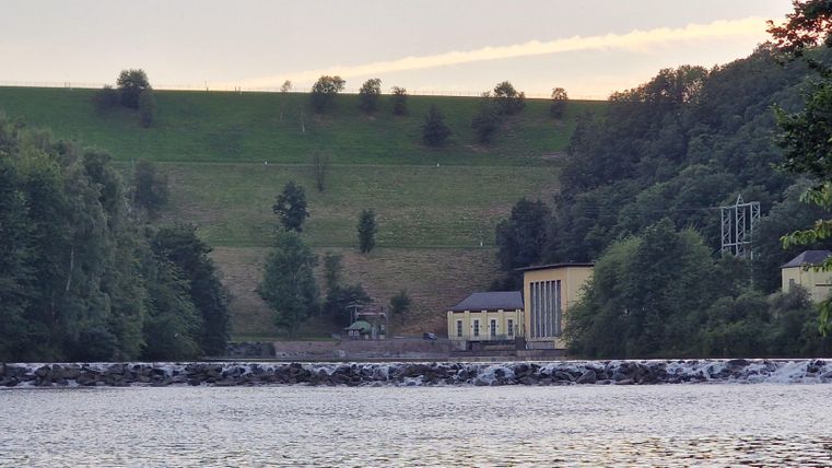 Ein ruhiger Fluss mit einer Mauer im Vordergrund. Im Hintergrund sind Hügel und Gebäude umgeben von Bäumen zu sehen.