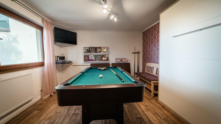 Ein Billardtisch steht in einem hellen Raum mit Holzfußboden. Im Hintergrund sind Regale und ein Fernseher sichtbar.