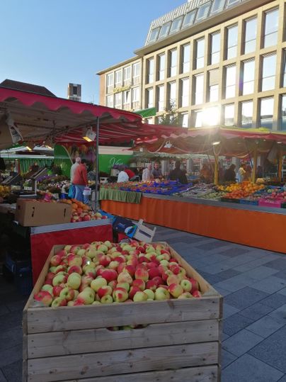 Ein lebhafter Markt mit frischen Äpfeln in einem Holzkorb. Im Hintergrund sind bunte Stände mit Obst und Gemüse zu sehen.