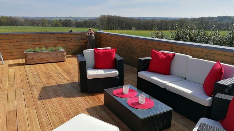 Eine gemütliche Terrasse mit modernen Sitzmöbeln und roten Kissen. Im Hintergrund erstreckt sich eine grüne Landschaft unter einem klaren Himmel.