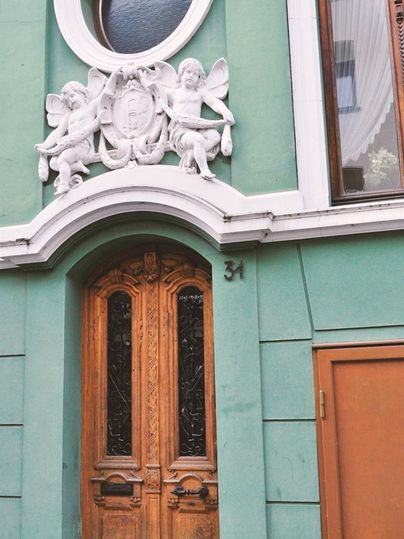 Eine grüne Hausfassade mit einem kunstvollen Relief von zwei Engeln über einer hölzernen Doppeltür. Die Tür hat Glasverzierungen und ist mit der Hausnummer 34 versehen.