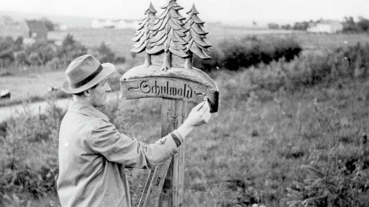 Ein Mann malt ein Schild mit der Aufschrift "Schlumath". Im Hintergrund sind grüne Wiesen und Bäume zu sehen.