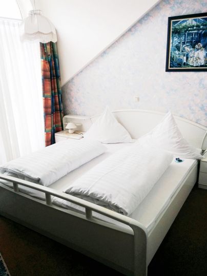 Ein gemütliches Schlafzimmer mit einem weißen Bett und zwei großen Kissen. Neben dem Fenster hängt ein farbenfrohes Vorhang und ein Bild ist an der Wand.