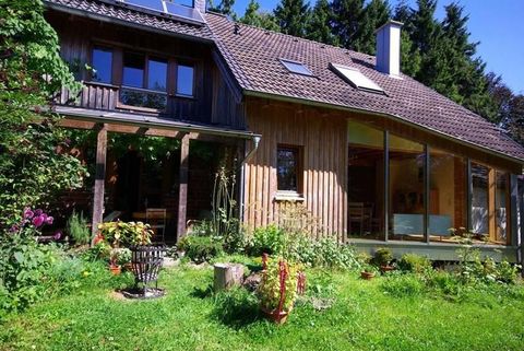 Ein modernes Holzhaus umgeben von einem üppigen Garten. Die Fassade hat große Fenster und der Bereich ist hell beleuchtet.