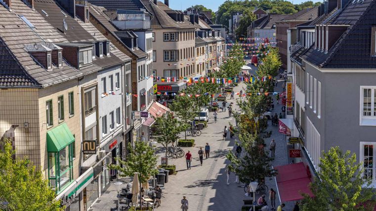 Eine belebte Einkaufsstraße mit Bäumen und vielen Menschen. Farbenfrohe Fahnen schmücken die Straße und laden zum Bummeln ein.