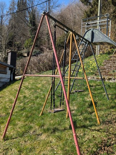 Ein Spielplatz mit einer Wippe und einer Rutsche in einem grünen Garten. Im Hintergrund sind Bäume und ein blauer Himmel zu sehen.