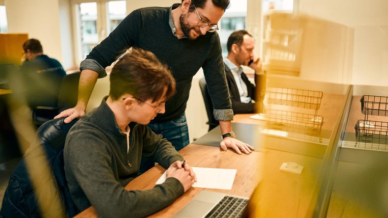 Zwei Männer sitzen an einem Schreibtisch und arbeiten zusammen. Einer gibt dem anderen Anweisungen, während ein weiterer Mann im Hintergrund beschäftigt ist.
