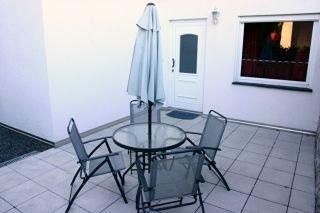 Ein gemütlicher Außenbereich mit einem Tisch und vier Stühlen. Ein Sonnenschirm steht auf der Terrasse vor einer weißen Wand.