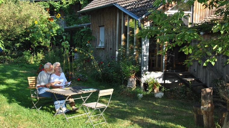 Ein älteres Paar sitzt gemütlich an einem Tisch im Garten und liest. Umgeben von Pflanzen und einem Holzhaus, strahlt die Szene Ruhe und Entspannung aus.