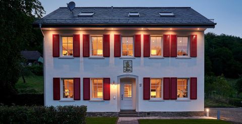 Ein modernes, zweistöckiges Haus mit weißen Wänden und roten Fensterläden. Die Fenster sind beleuchtet und es gibt einen schmalen Weg zur Eingangstür.