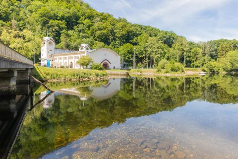 Ein ruhiger See mit klarer Reflexion der umliegenden Natur. Im Hintergrund ist ein historisches Gebäude und üppiges Grün zu sehen.