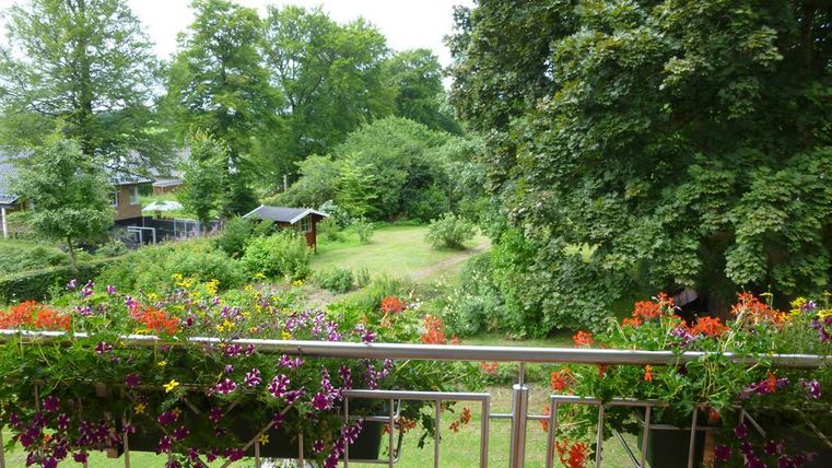 Ein schöner Ausblick vom Balkon auf einen grünen Garten mit bunten Blumen. Im Hintergrund sind Bäume und ein kleiner Schuppen zu sehen.