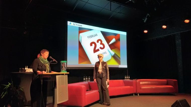 Eine Präsentation auf einer Bühne mit einem Kalenderblatt, das den 23. Februar zeigt. Zwei Personen stehen auf der Bühne vor roten Sofas.