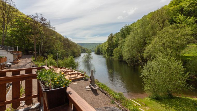 Ein ruhiger Fluss, umgeben von üppigem Grün und Bäumen. Im Vordergrund sind Holzterrassen und Blumenkästen zu sehen.
