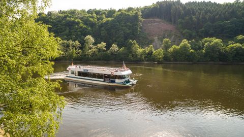 Ein Boot fährt auf einem ruhigen Fluss, umgeben von grünen Bäumen. Die Landschaft im Hintergrund zeigt sanfte Hügel und einen blauen Himmel.