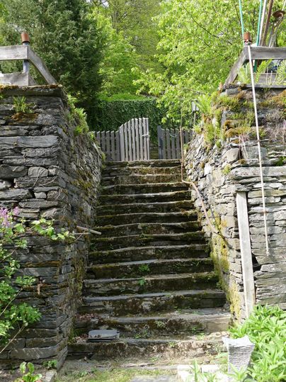 Eine steinerne Treppe führt zu einem offenen Garten. Moos bedeckt einige Stufen, und im Hintergrund ist ein Holztor sichtbar.