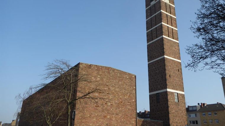 Ein modernes Gebäude aus rotem Stein mit einem markanten, hohen Turm. Der Himmel ist klar und die Umgebung ist belebt.