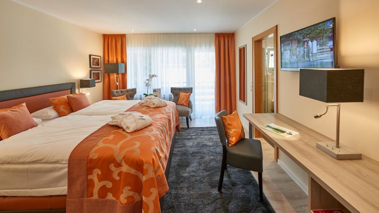Ein gemütliches Hotelzimmer mit zwei Betten, orangefarbener Bettwäsche und modernen Möbeln. Große Fenster sorgen für viel Licht und eine angenehme Atmosphäre.