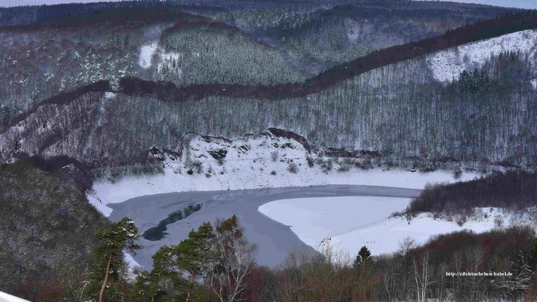 Eine winterliche Landschaft mit einem zugefrorenen Fluss und schneebedeckten Bäumen. Die Hügel im Hintergrund sind mit Fichten bewachsen.