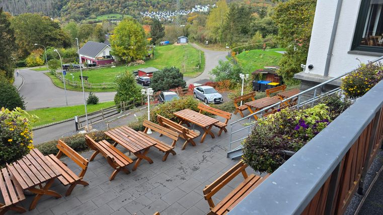 Eine gemütliche Terrasse mit Holztischen und Bänken. Im Hintergrund erstreckt sich eine grüne Landschaft mit saftigen Bäumen und sanften Hügeln.