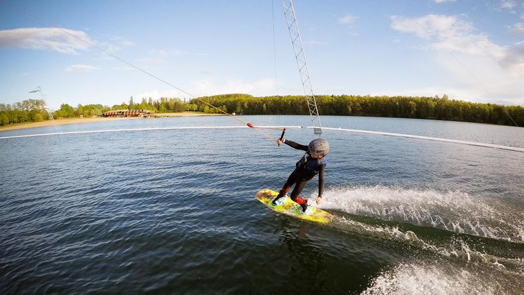 Ein Wassersportler fährt auf einem Wakeboard über ruhiges Wasser. Im Hintergrund sind Bäume und ein blauer Himmel sichtbar.