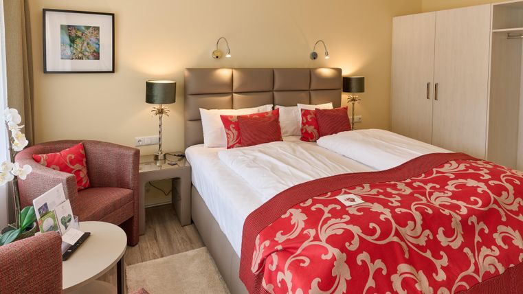 Ein elegantes Schlafzimmer mit einem großen Bett, roten Kissen und einer gemütlichen Sitzecke. Die Wände sind in sanften Tönen gehalten, und es gibt eine harmonische Beleuchtung.