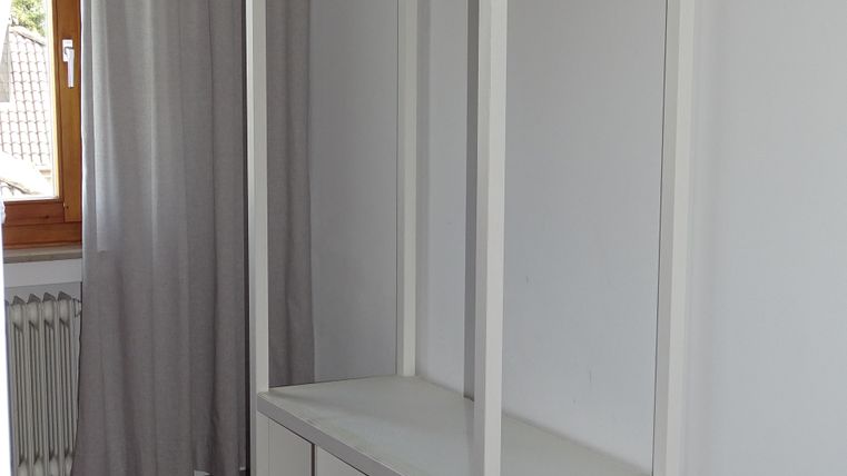 Ein einfacher, weißer Kleiderständer mit Holzkleiderbügeln steht an der Wand. Darunter befindet sich eine Ablage mit offenen Fächern.