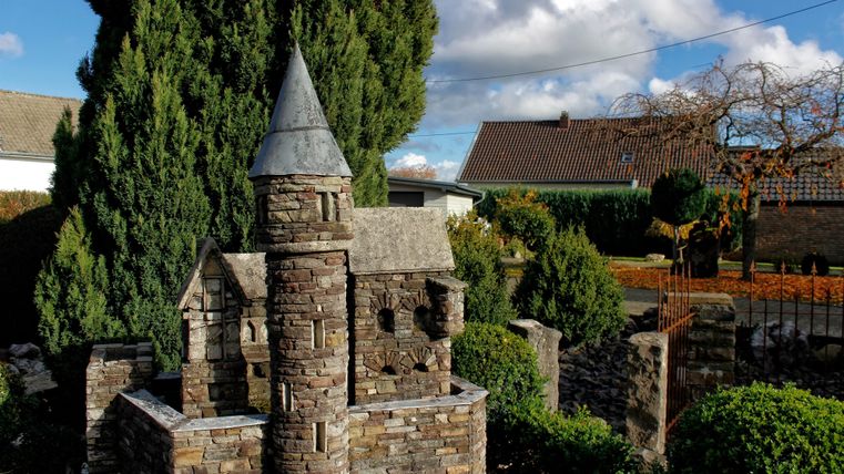 Ein wunderschönes Steinschloss mit einem Turm in einem Garten. Im Hintergrund sind grüne Bäume und ein Haus sichtbar.