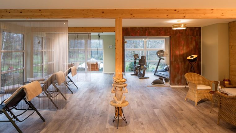 Ein moderner Raum mit Holzdetails und großen Fenstern. Es gibt Liegen und Fitnessgeräte, sowie einen gemütlichen Bereich zum Entspannen.