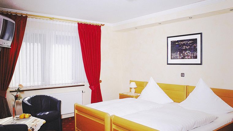 Ein gemütliches Hotelzimmer mit zwei Betten, einem Fernseher und einem Sessel. Rote Vorhänge und ein Wandbild sorgen für eine einladende Atmosphäre.