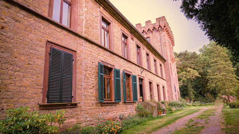 Ein historisches Gebäude mit Steinfassade und grünen Fensterläden. Der Weg führt durch einen Garten mit üppiger Vegetation.