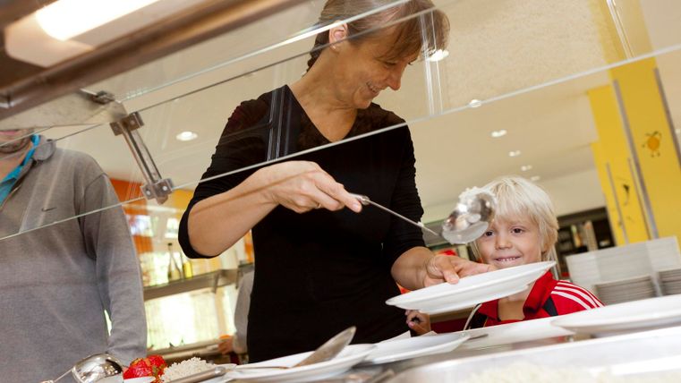 Eine Frau serviert Essen an einem Tisch in einer Kantine. Ein glückliches Kind schaut lächelnd zu ihr auf.